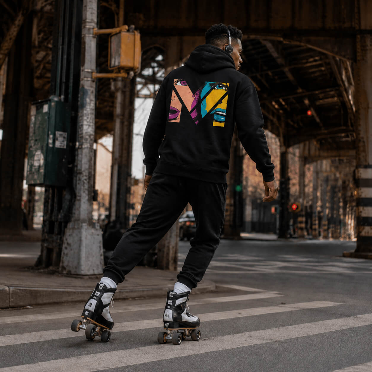 Collection Impact & Nostalgie, homme noir en rollers sous le métro aérien de New York, hoodie noir avec design pop art coloré au dos, ambiance urbaine rétro Mindset Ambition