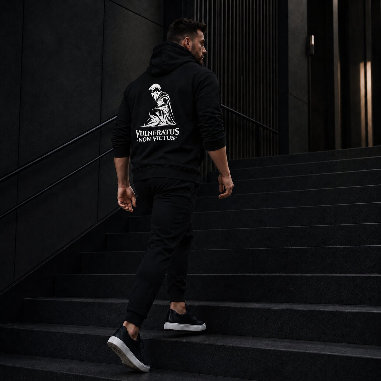 Homme en hoodie noir Vulneratus Non Victus montant un escalier sombre Honneur & Discipline Mindset Ambition